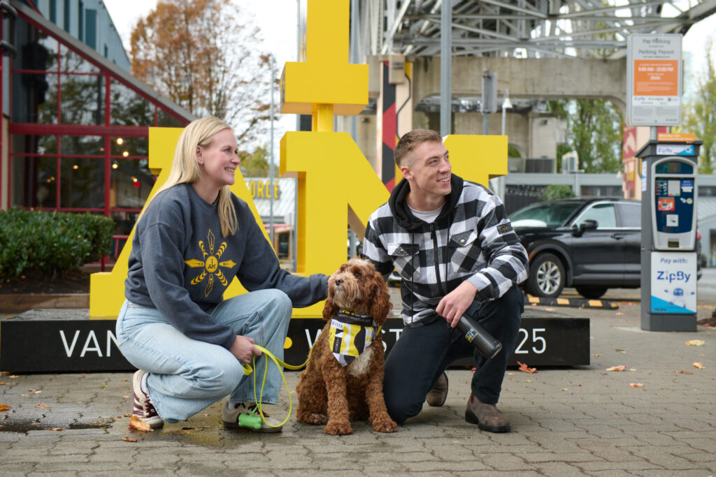 A cheerful outdoor scene featuring a couple with their dog in front of the yellow 'I AM' installation for the Invictus Games Vancouver Whistler 2025. The woman wears a grey sweatshirt with an Invictus Games design, kneeling and holding the dog's leash, while the man in a checkered jacket crouches beside them, holding a branded water bottle. The dog, sporting an Invictus Games bandana, gazes upward, adding a touch of warmth and community spirit to the image.